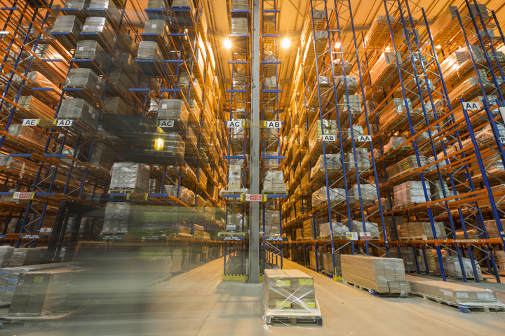 Large distribution warehouse with multi‑level pallet racking, stacked inventory, and a forklift moving pallets along a narrow aisle.