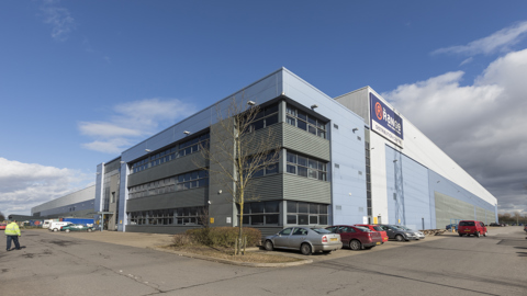 Large modern industrial warehouse with branded signage and adjacent parking area under a clear blue sky.