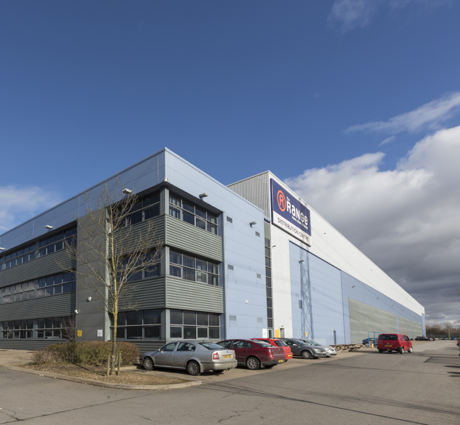 Large modern industrial warehouse with branded signage and adjacent parking area under a clear blue sky.