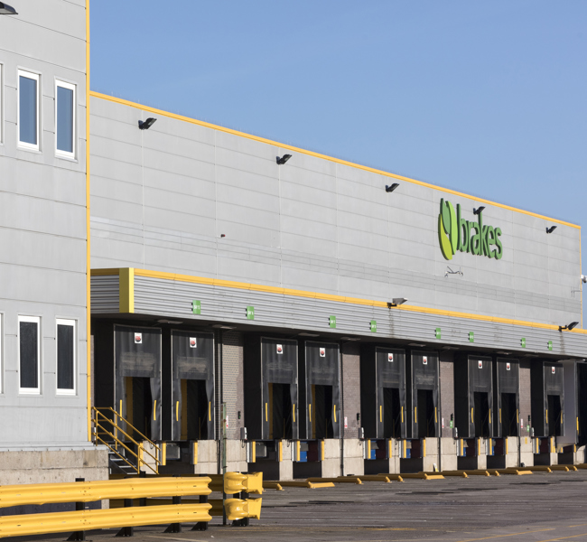 Large industrial warehouse with multiple loading docks and Brakes signage, modern UK logistics facility